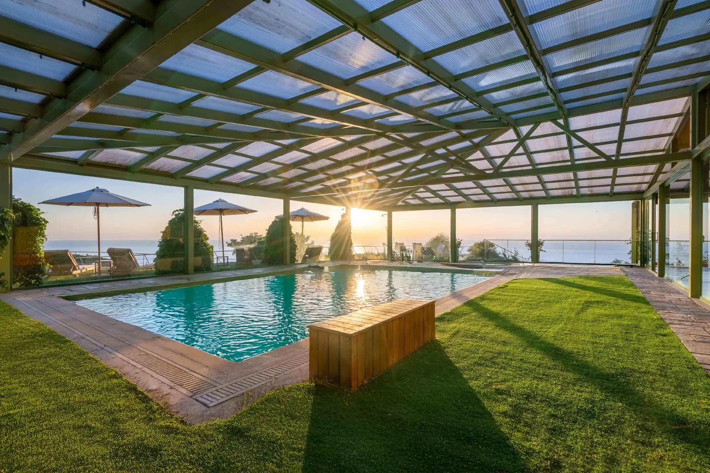 Outdoor pool with a wooden bench, green grass, and ocean view under a glass roof at sunset.