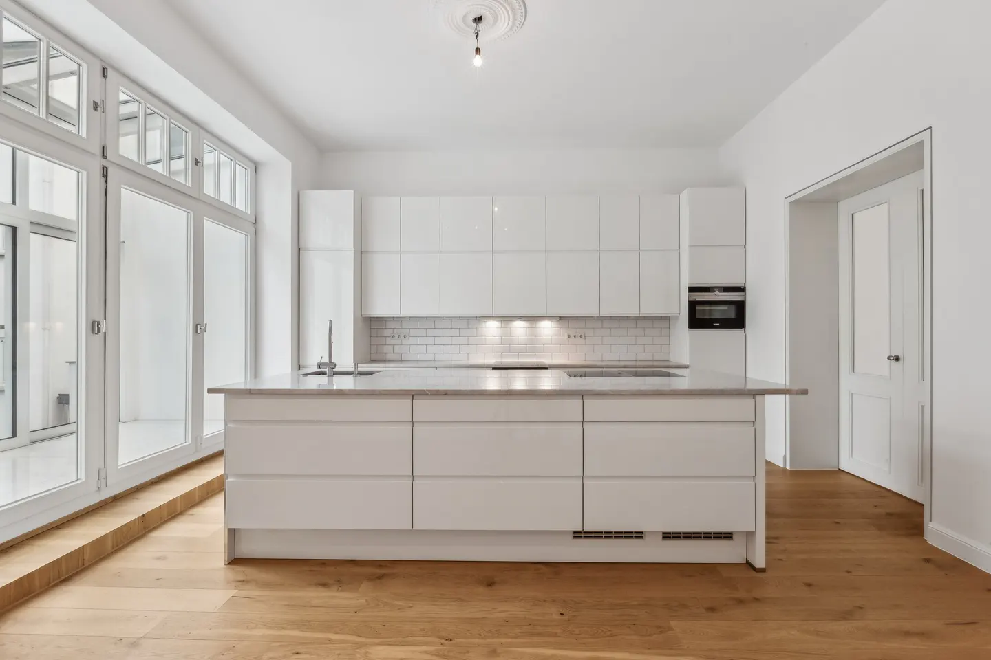 Bright, modern kitchen with white cabinets, marble island, and wood floors. Large windows provide natural light.