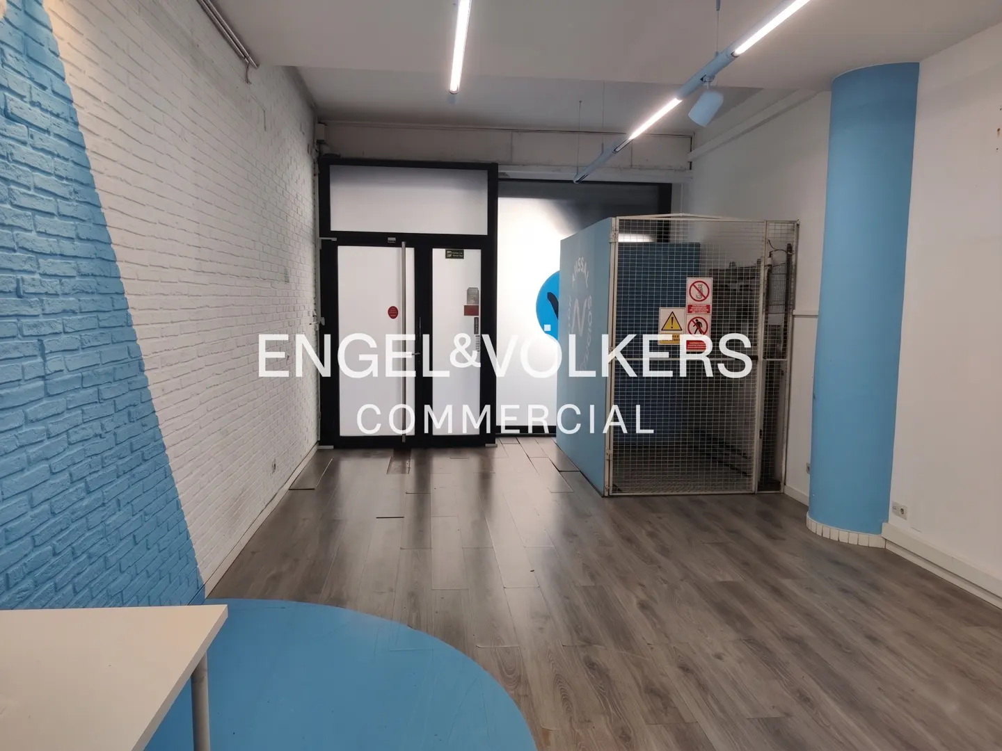 An empty commercial space with wood floors, white brick walls with blue accents, and a black-framed glass door.