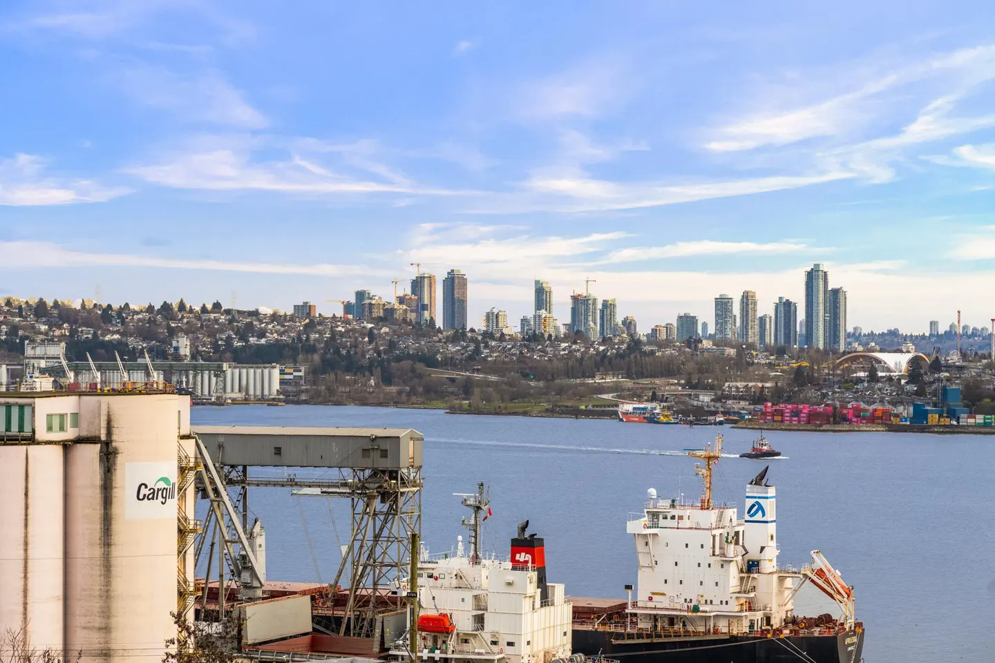 Cityscape view of Vancouver, BC, with cargo ships in the foreground and a Cargill grain elevator on the left. Blue sky with wispy clouds.