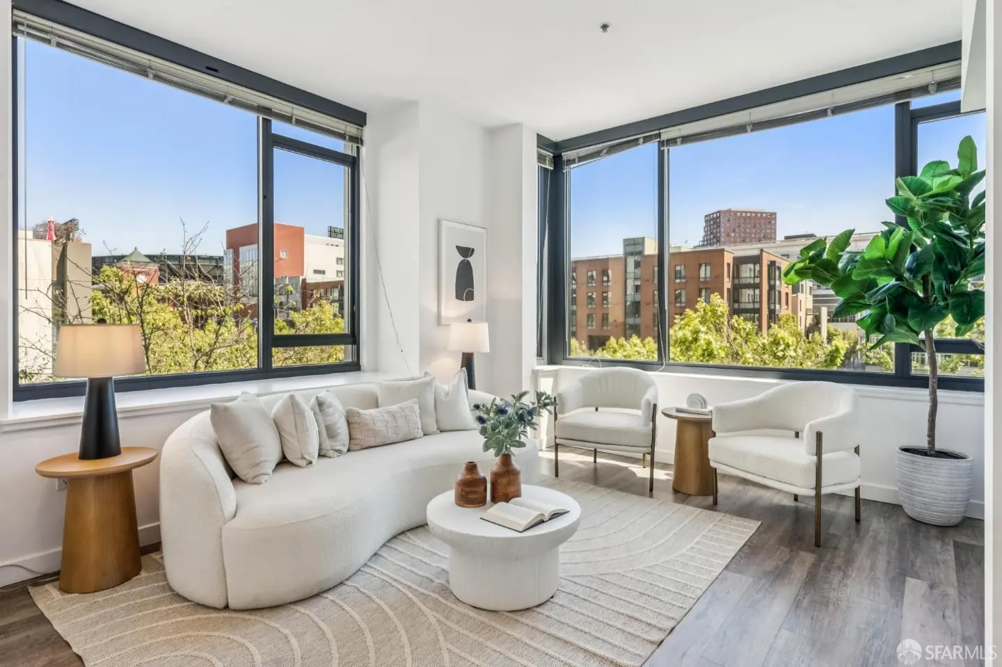 Bright living room with a curved white sofa, two chairs, and large windows overlooking a city view. A potted plant sits near the window.