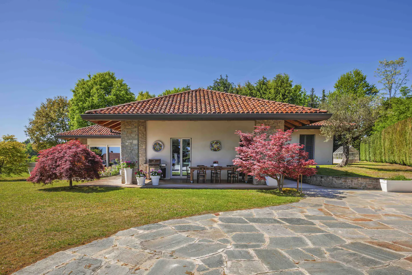 A single-story house with a red tile roof, stone accents, and a patio with a wooden table and chairs. A red Japanese maple tree is in the front yard.