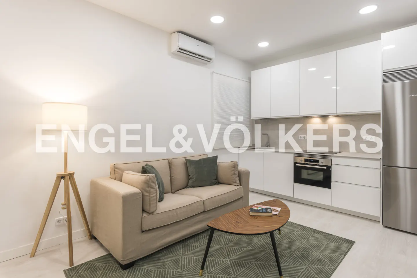 Bright, modern apartment interior with a beige sofa, green rug, and white kitchen cabinets. A wooden floor lamp stands near the sofa.