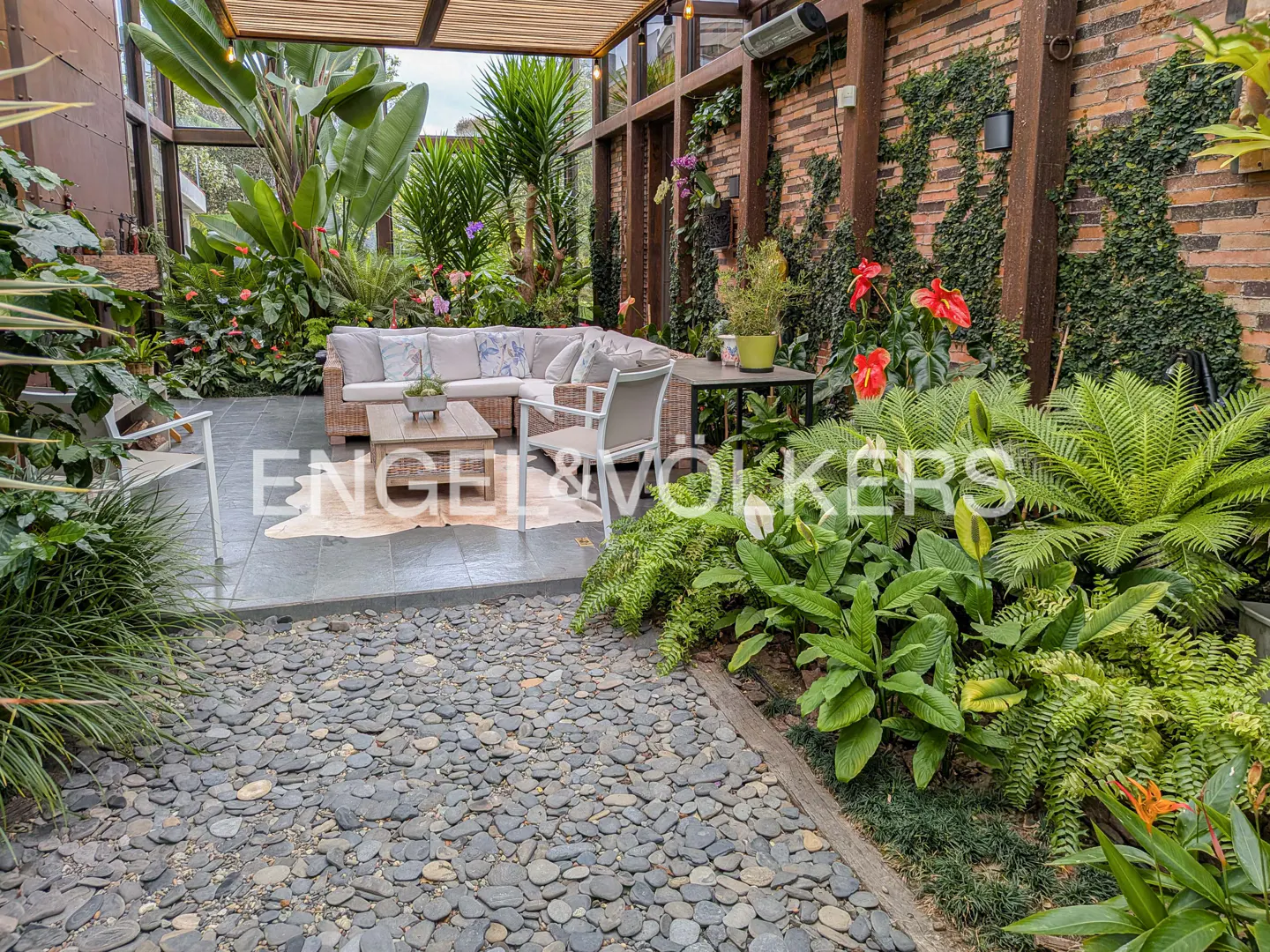 Outdoor patio with wicker furniture, lush greenery, and a stone walkway. A brick wall with climbing vines adds a rustic touch.
