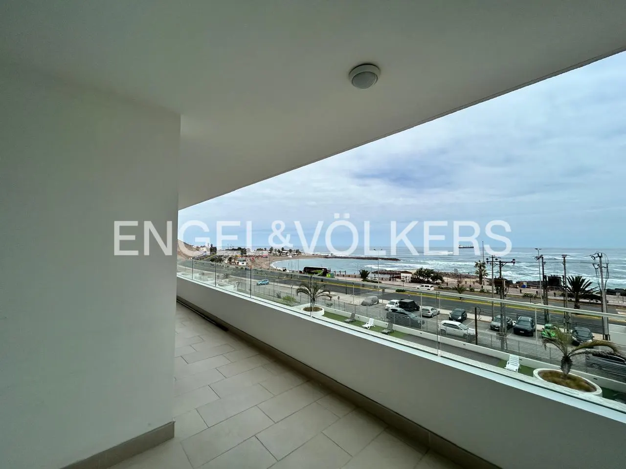Balcony view of ocean and city street. White walls, glass railing, and Engel & Völkers logo.