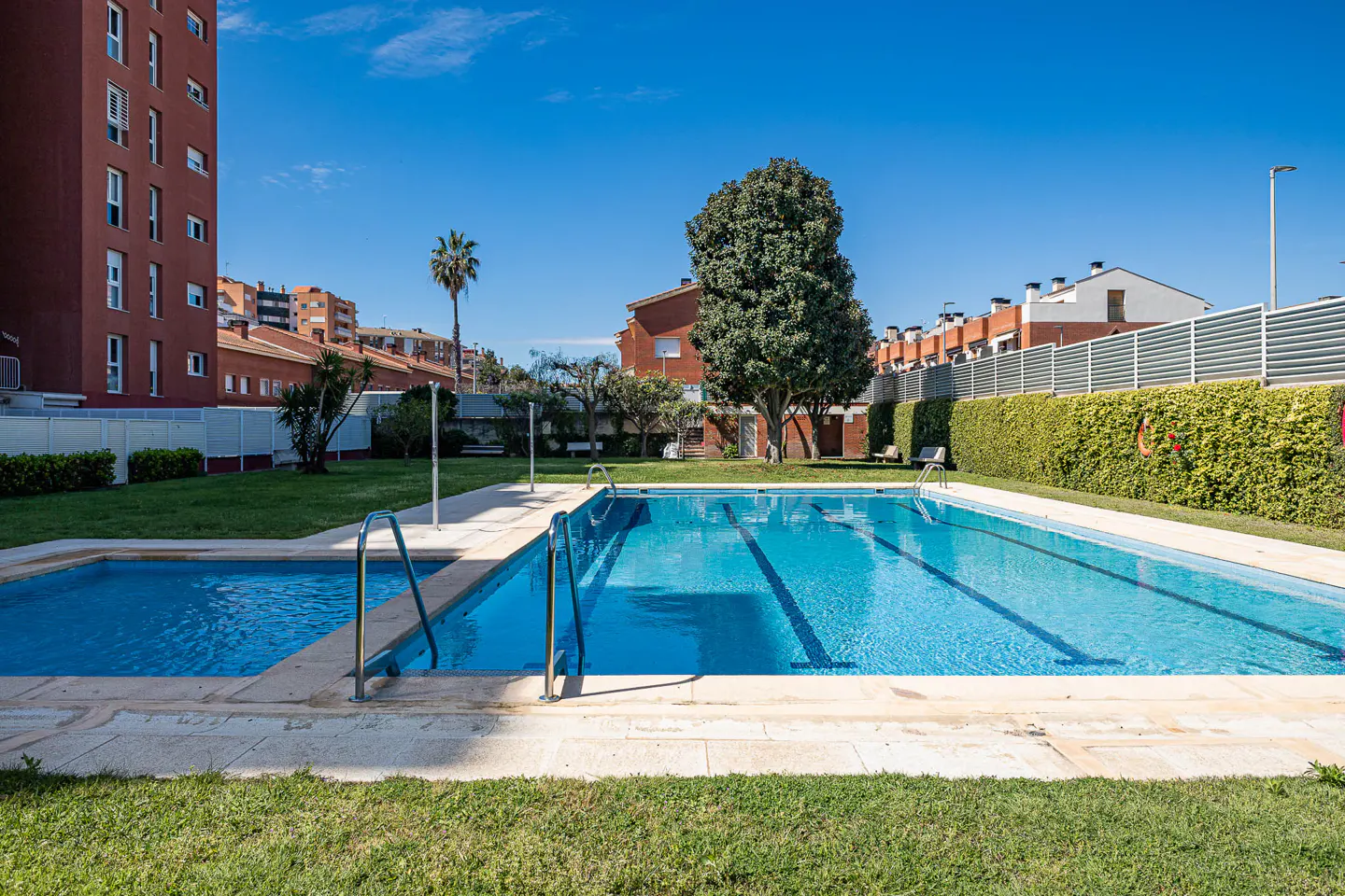 Outdoor pool with blue water and metal ladders, surrounded by green grass and hedges, with buildings in the background under a clear blue sky.