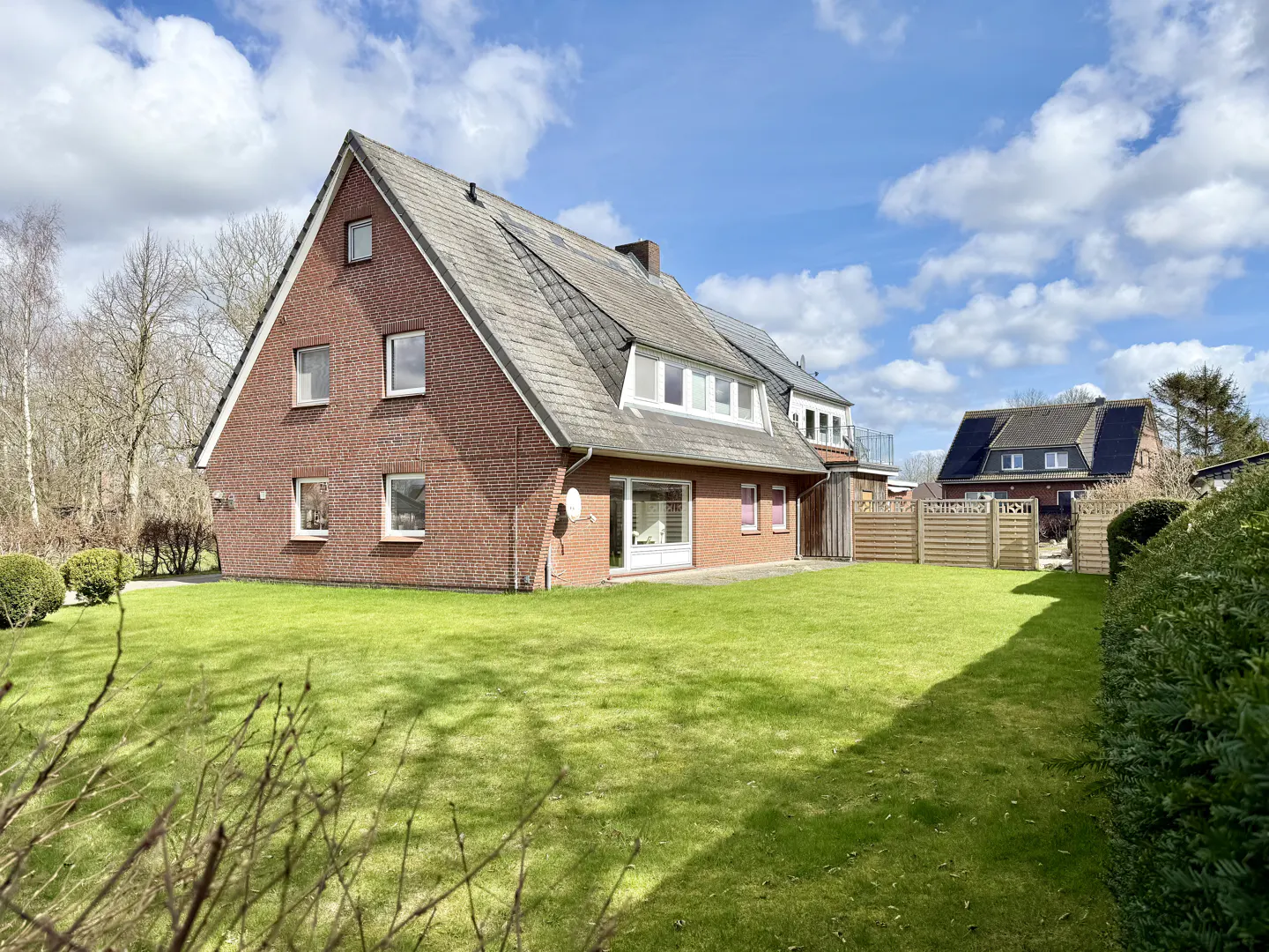 A two-story brick house with a gray roof sits on a green lawn under a blue sky with white clouds.