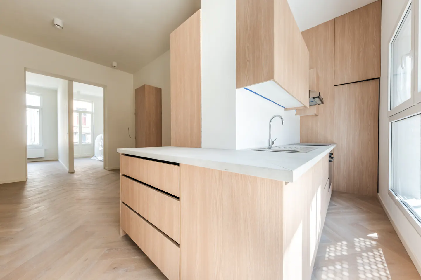 A bright, modern kitchen with light wood cabinets, a white countertop, and a stainless steel faucet. A herringbone wood floor extends into an adjacent room.