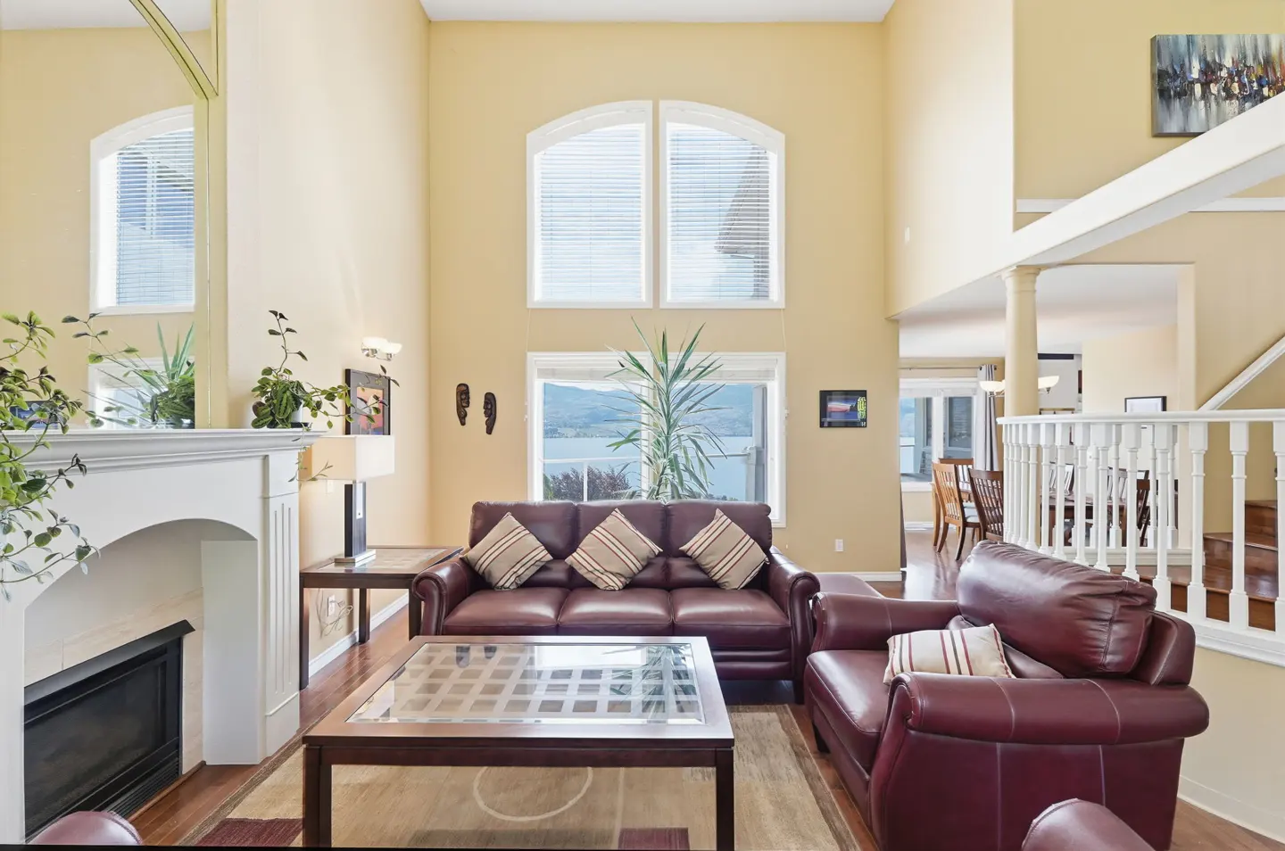 Spacious living room with high ceilings, burgundy leather sofas, a glass coffee table, fireplace, and lake view windows.