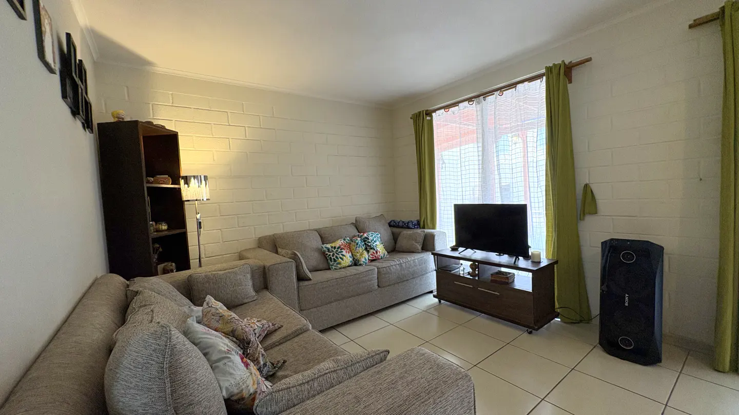 Living room with gray sectional sofa, floral pillows, TV on a brown stand, and a black speaker. Green curtains frame a window.