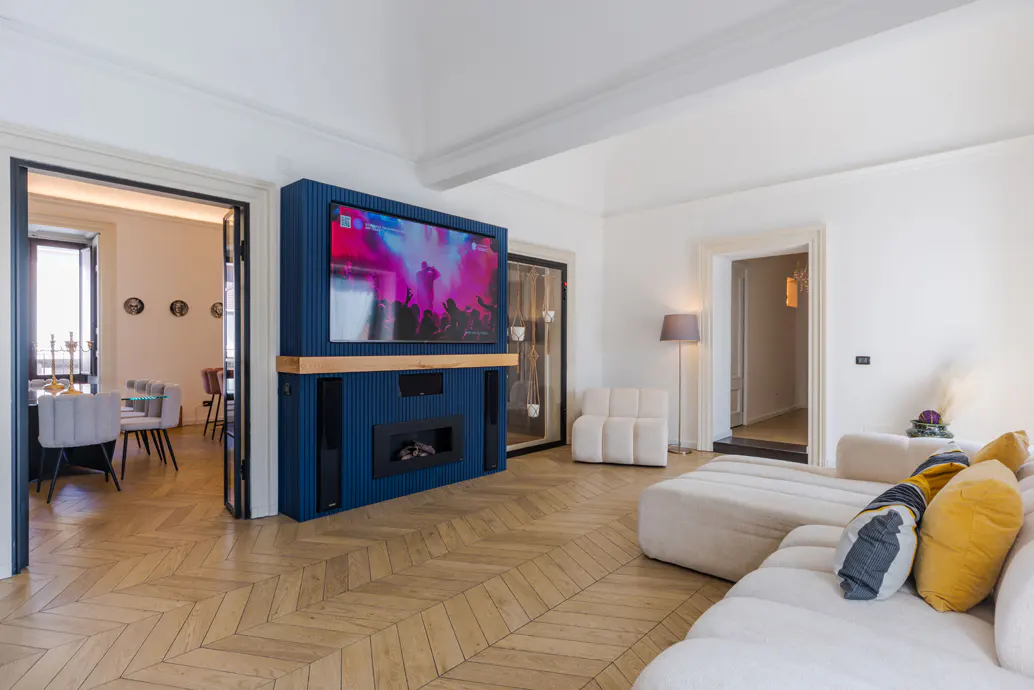 Bright living room with herringbone wood floors, white walls, and a blue entertainment center with a TV and fireplace. A white sectional sofa is in the foreground.
