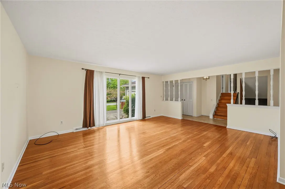 Bright living room with hardwood floors, cream walls, and a large sliding glass door with brown curtains. Stairs are visible in the background.