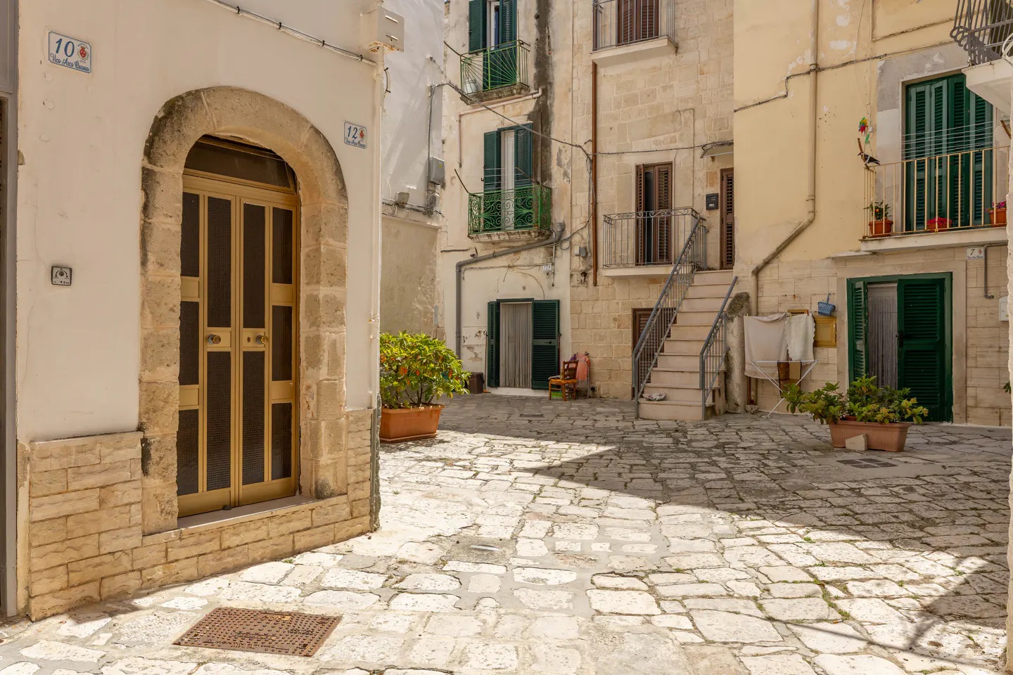 A sunny, stone-paved courtyard in an Italian village with historic buildings, green shutters, balconies, and potted plants.