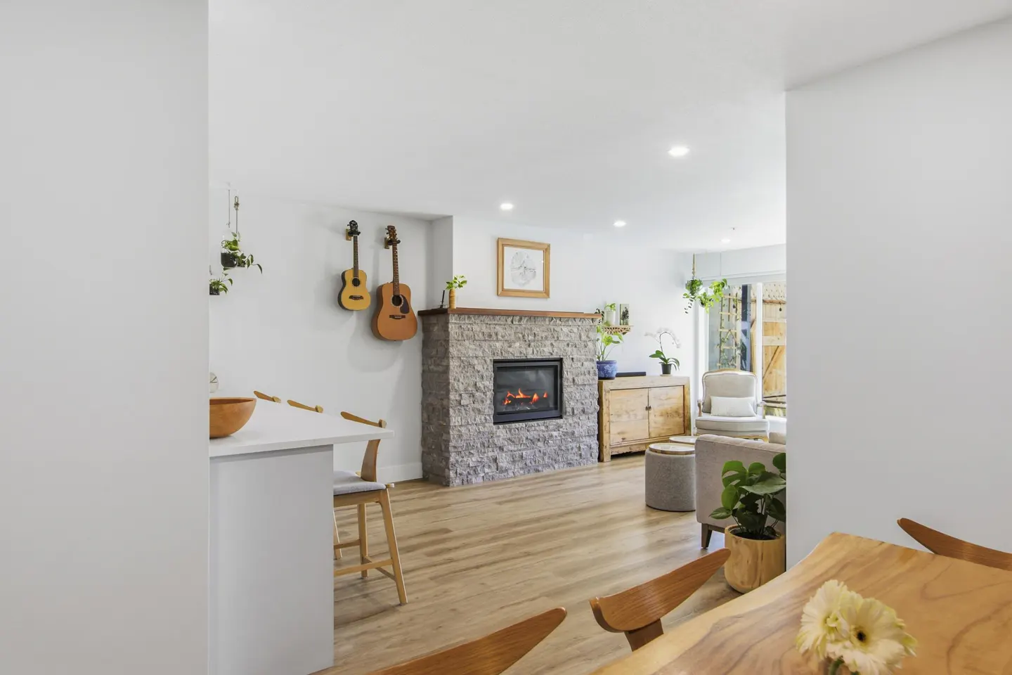 Bright living room with a stone fireplace, hardwood floors, and white walls. Two guitars hang on the wall. A wooden table and chairs are in the foreground.
