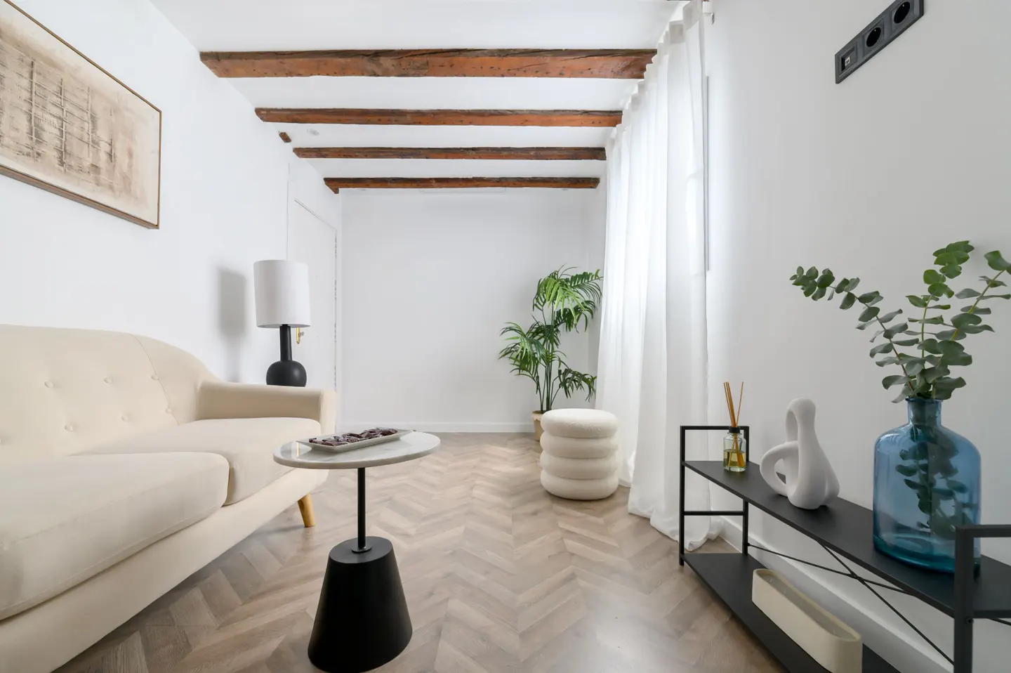 Bright living room with white walls, wood beams, and herringbone floors. A cream sofa, black table, and plants add warmth to the space.