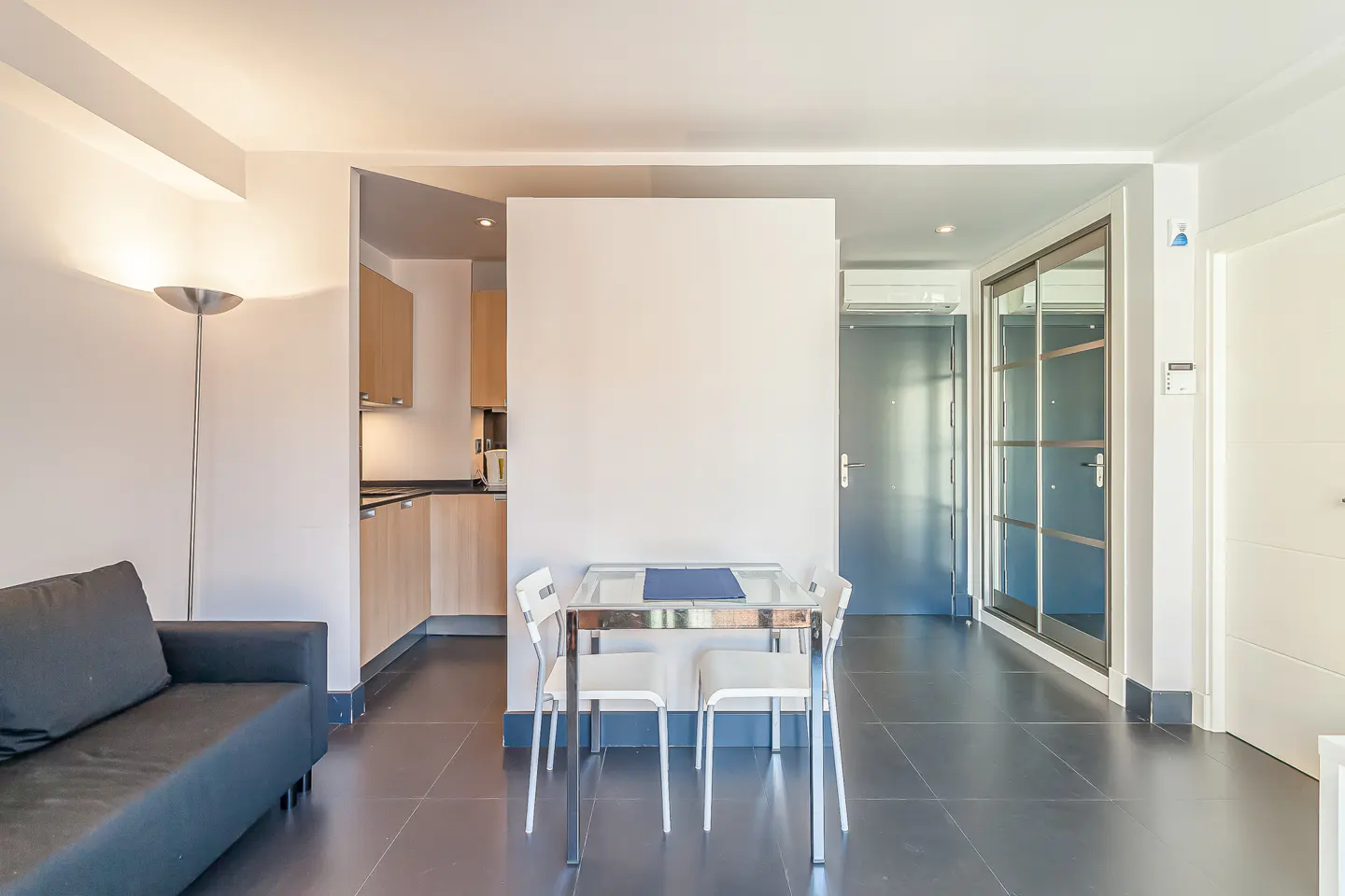 A modern apartment interior with a black sofa, dining table with four chairs, and a glimpse of the kitchen.