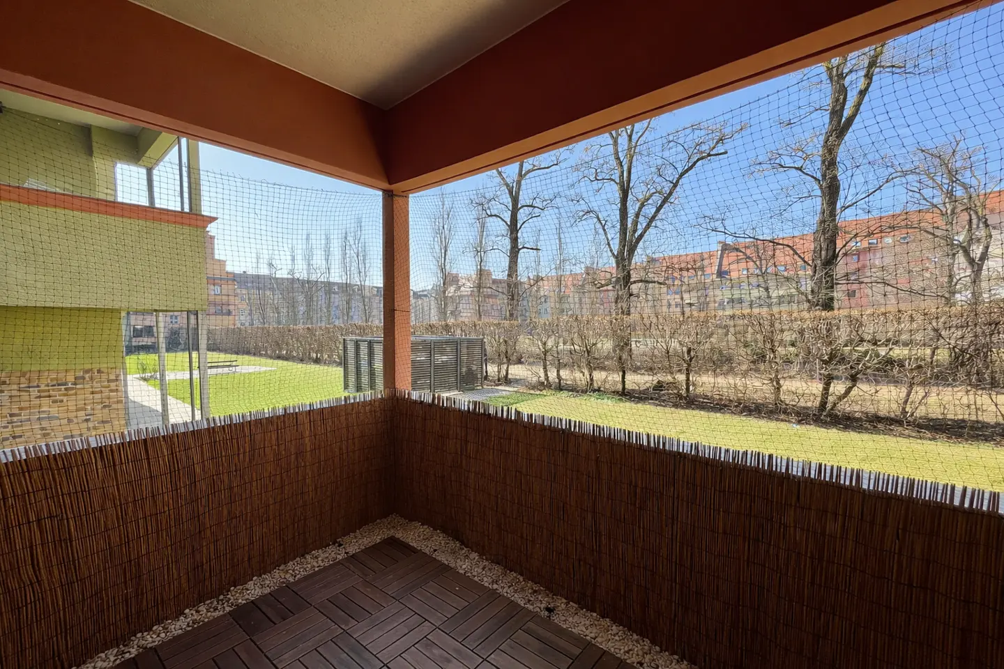 A balcony with a brown bamboo fence, a net, and a view of a green lawn and trees.