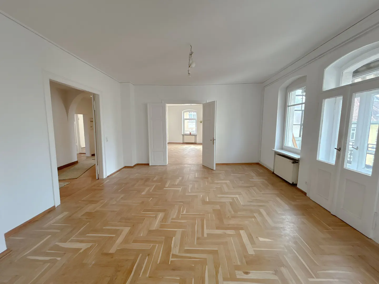 Bright, empty room with herringbone wood floors, white walls, and multiple doorways and windows.