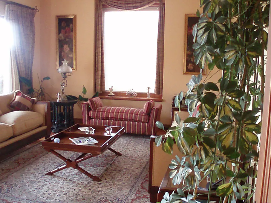 A living room with a beige sofa, striped bench, wooden table, and a large window. Paintings adorn the walls.