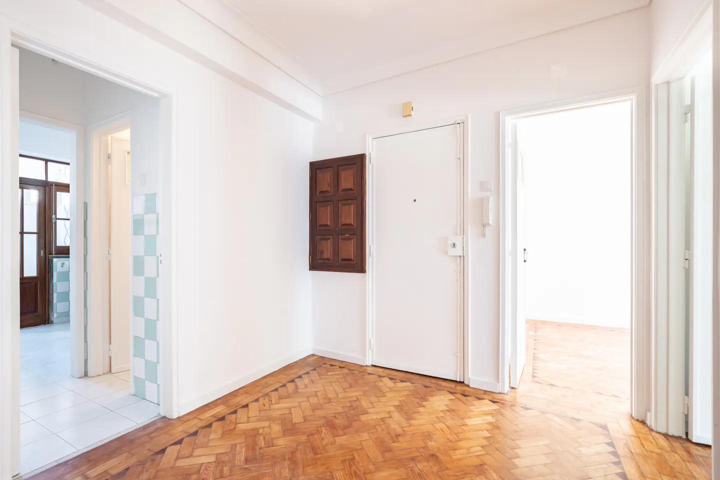 Bright hallway with herringbone wood floors, white walls, a white entry door, and doorways leading to other rooms.