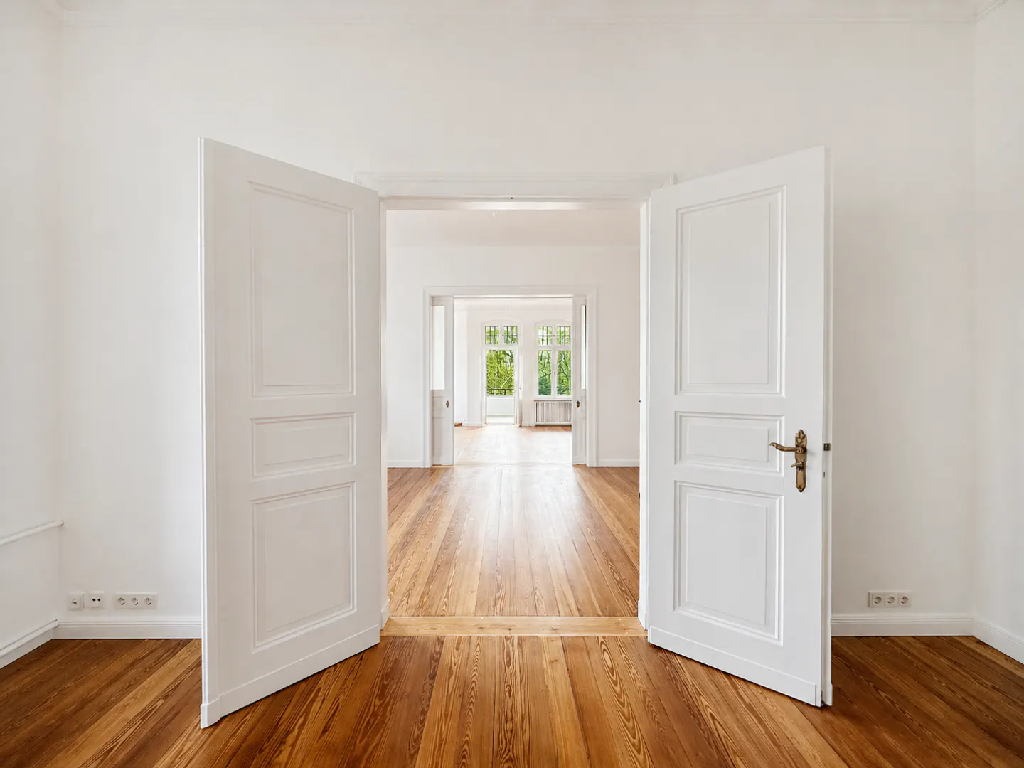 Open white double doors reveal a bright, empty room with hardwood floors and white walls, leading to another room with windows.