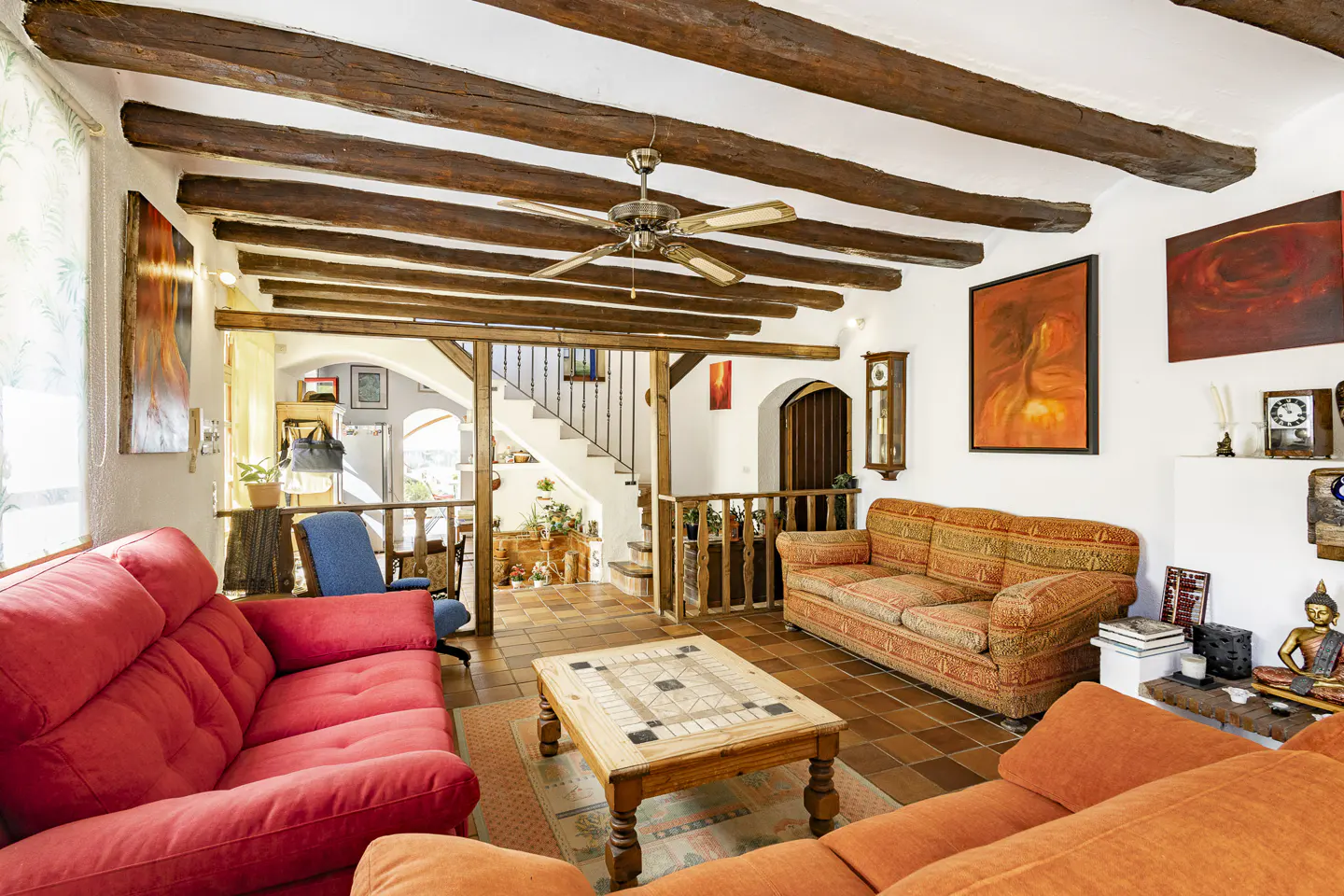 Living room with exposed wooden ceiling beams, a red sofa, orange sofa, wooden coffee table, and stairs in the background.
