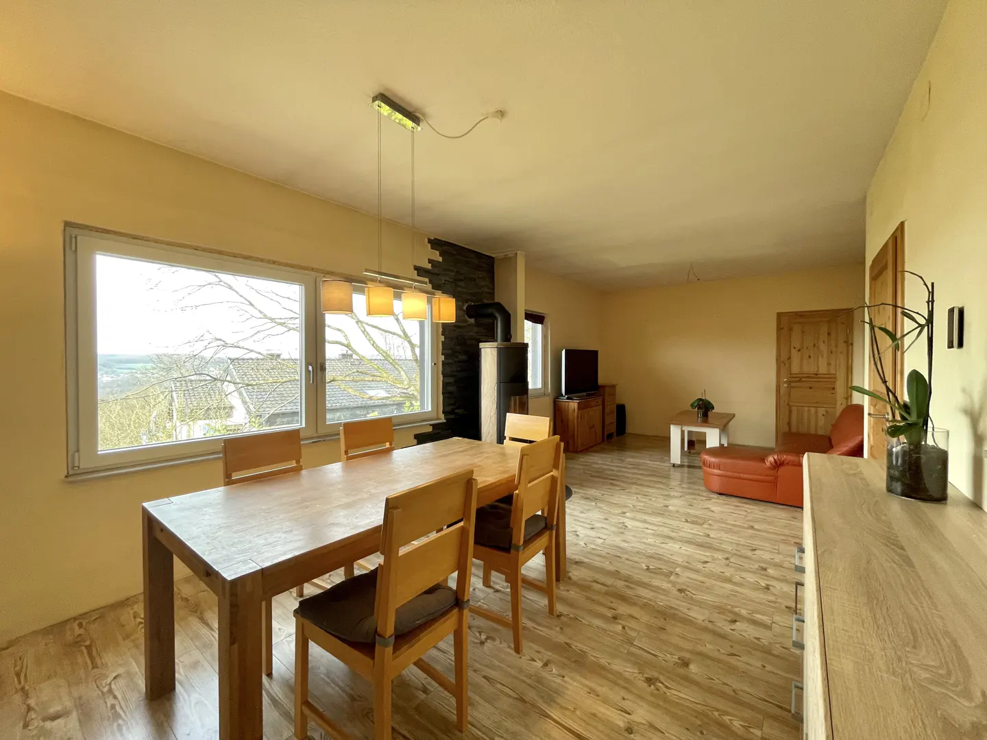 A bright, open-plan living and dining area with a wood dining table, chairs, and a large window with a view. A fireplace and seating area are visible in the background.