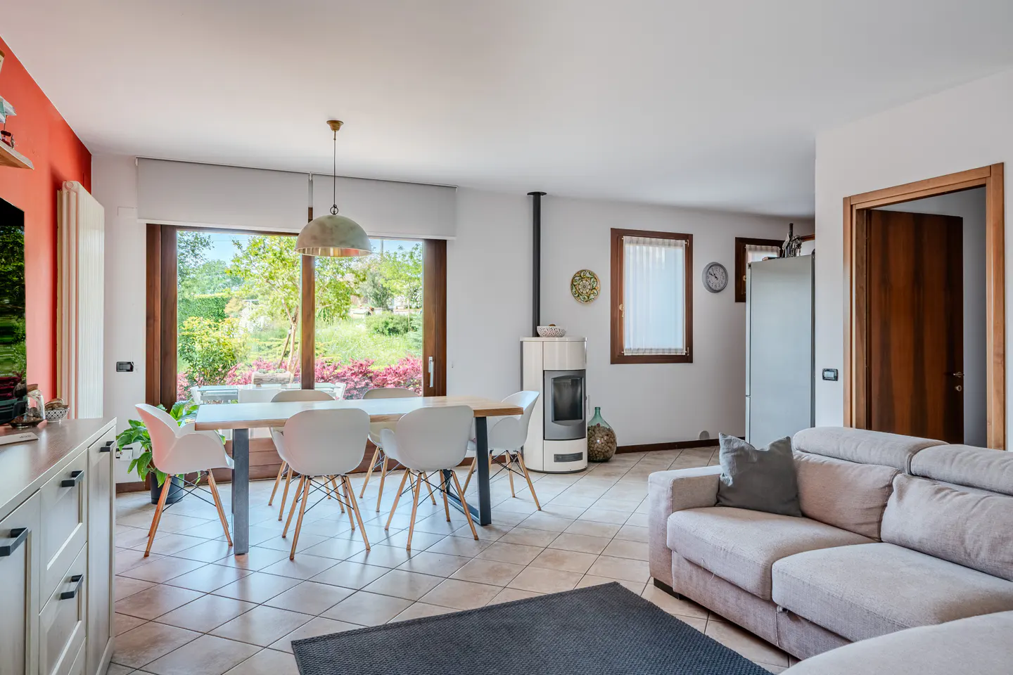 Bright living room with a dining table, white chairs, a beige sofa, and a wood-burning stove. Large window overlooks a lush garden.