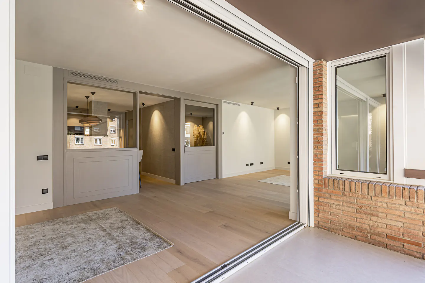 Open-concept living space with light wood floors, white walls, and sliding glass doors to a brick-walled patio. Kitchen visible through a glass partition.