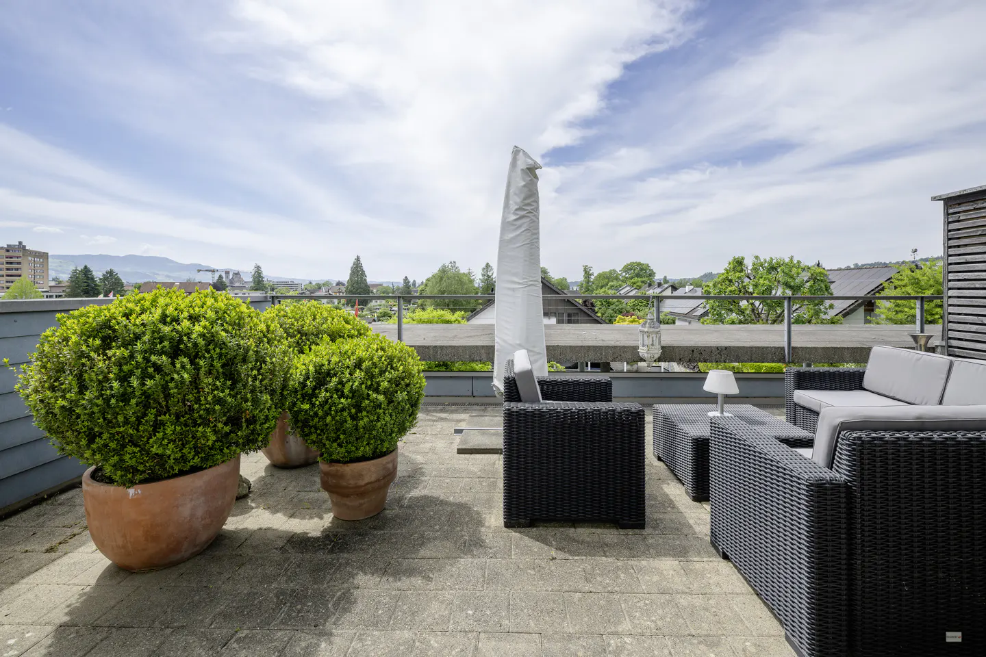 Rooftop patio with wicker furniture, potted green shrubs, and a covered umbrella. Cityscape and blue sky in the background.