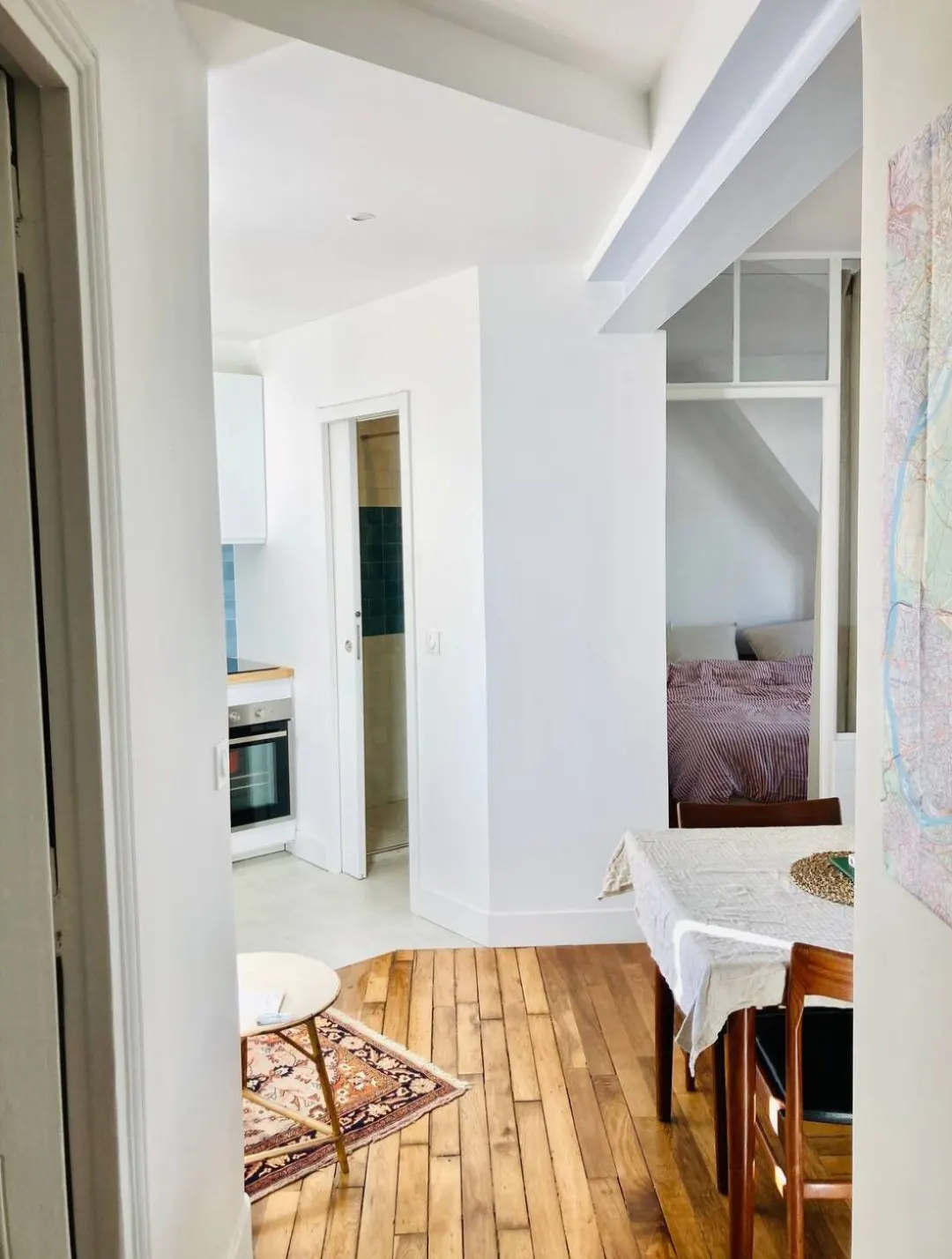 Bright apartment interior with wood floors, white walls, and a glimpse of a bedroom. A dining table with a white cloth is visible.