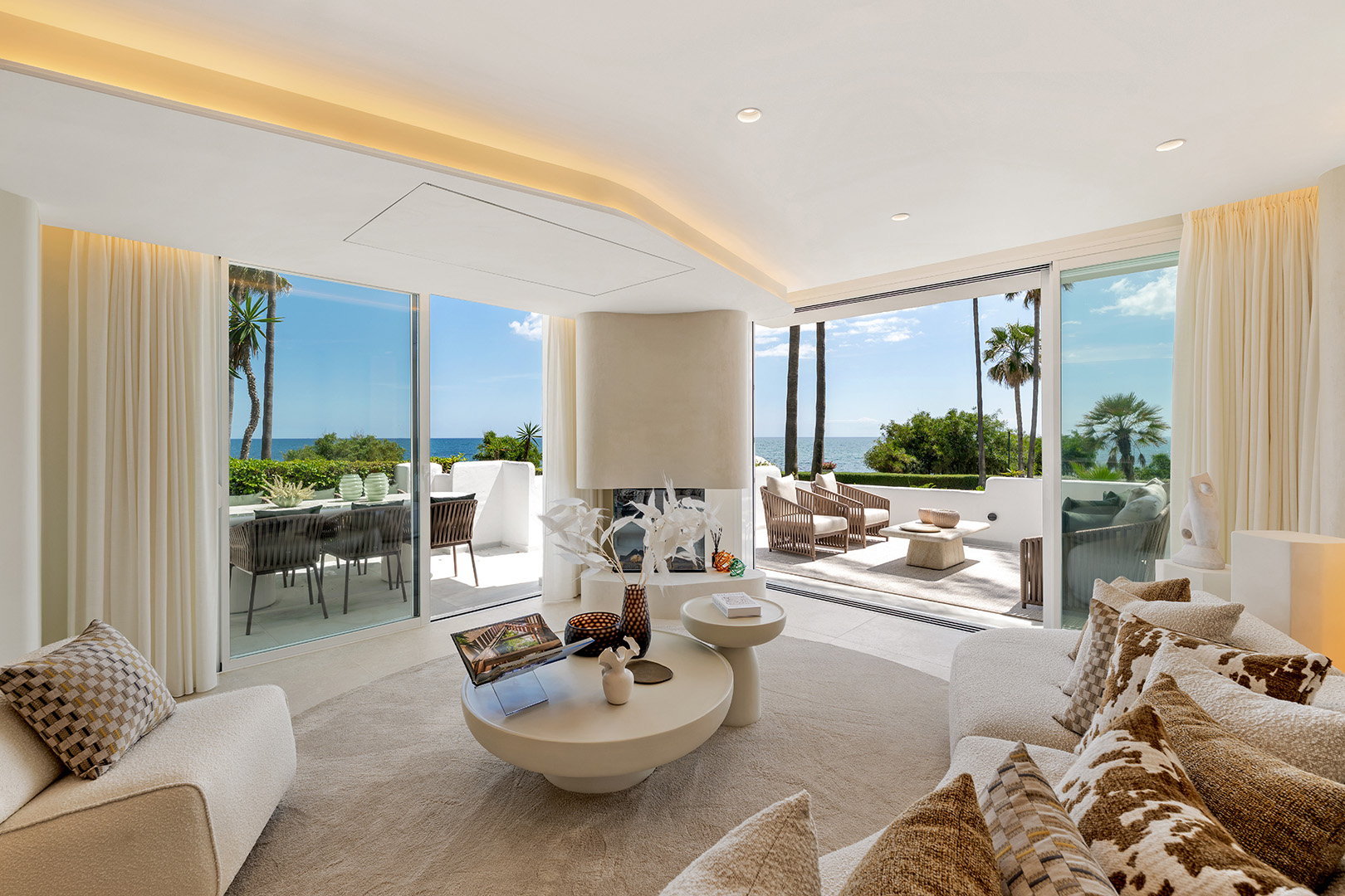Modern living room with cream furniture, a central fireplace, and large glass doors opening to a sunny ocean-view terrace.
