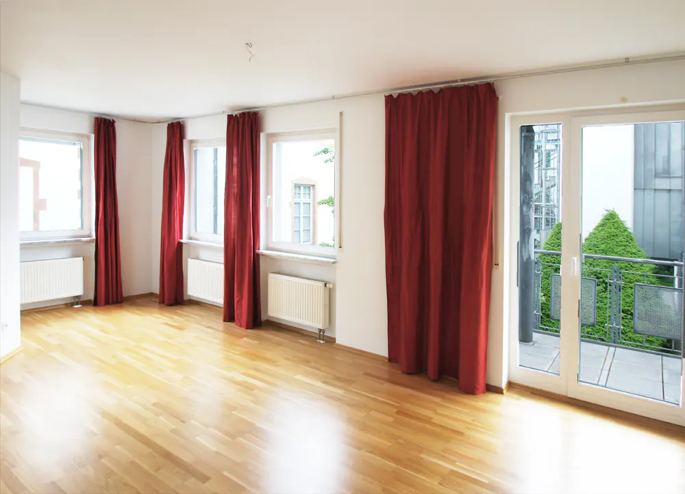 Empty room with wood floors, white walls, and red curtains. Windows and a glass door lead to a balcony with greenery.