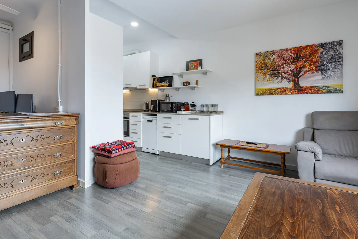 Open-concept living space with gray wood floors, white kitchen cabinets, and a brown wooden dresser. A colorful tree painting hangs on the wall.