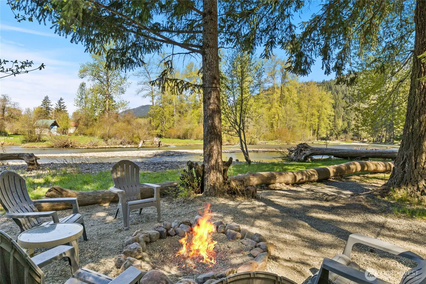 Outdoor seating area with a fire pit surrounded by gray Adirondack chairs, a river, and trees.