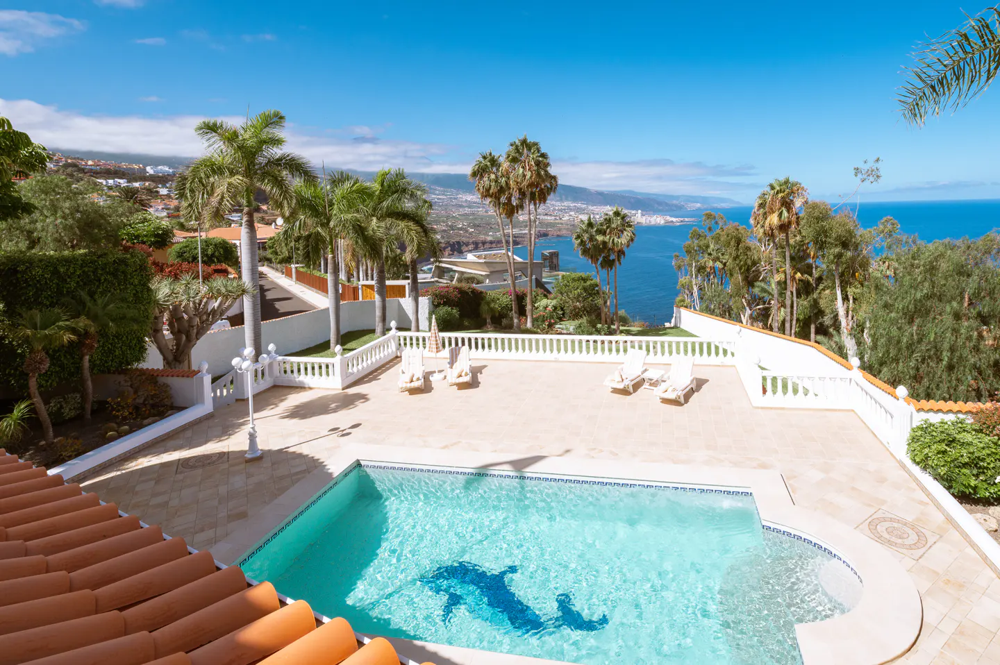 Aerial view of a pool with a dolphin mosaic, surrounded by lounge chairs, palm trees, and a blue ocean backdrop.