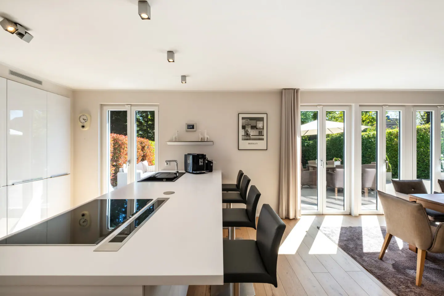 Bright, modern kitchen with a white island, black bar stools, and floor-to-ceiling windows overlooking a green patio.