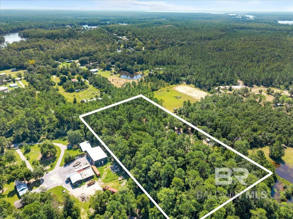 Aerial view of a wooded lot outlined in white, surrounded by forest, nearby buildings, and a small pond.