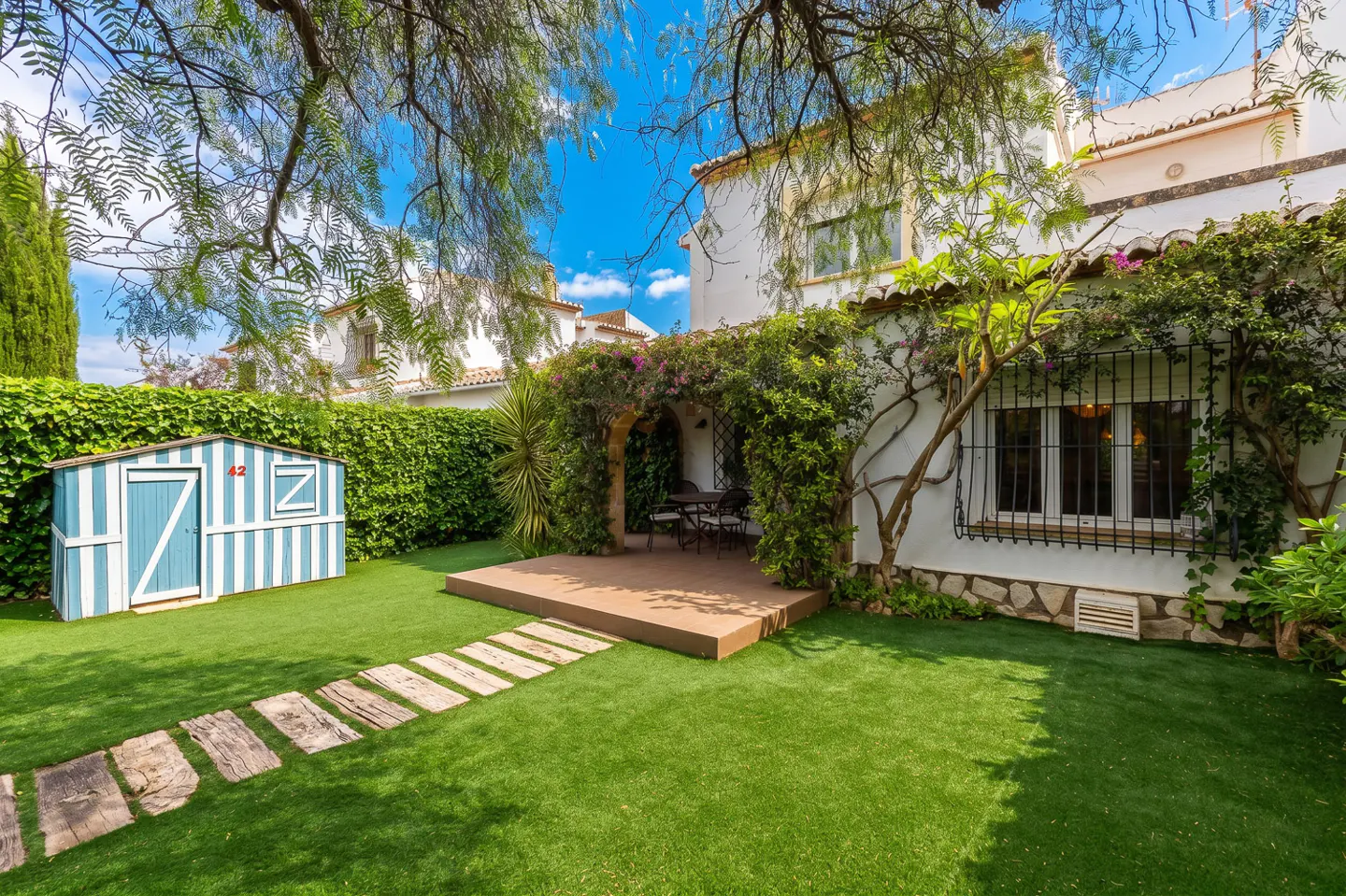A backyard with green grass, a blue and white shed, and a white house with vines.