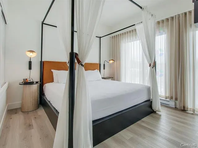 Bedroom with a black canopy bed, white linens, and tan headboard. Sheer white curtains frame the bed and windows. Wood floors.