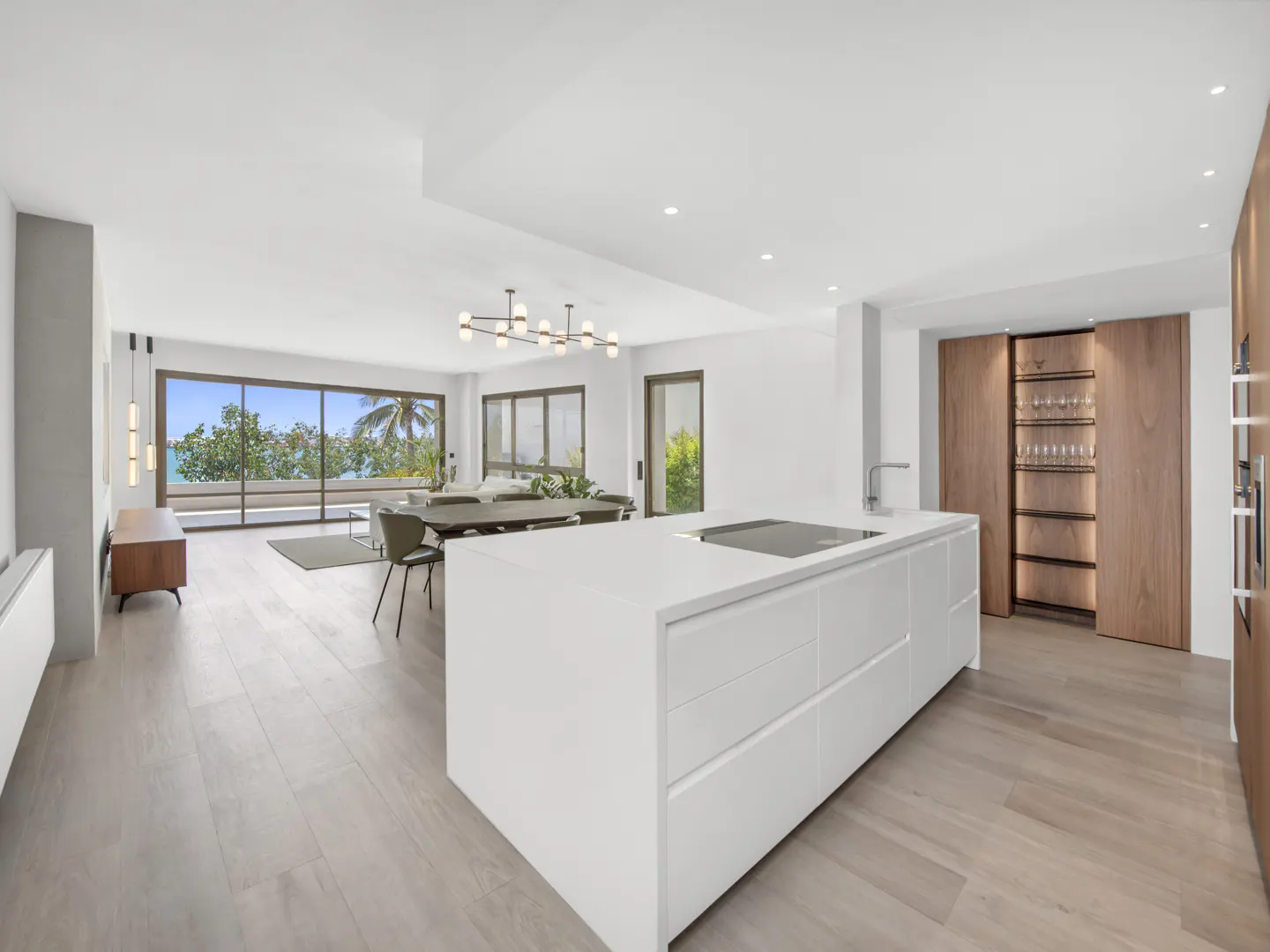 Bright, modern kitchen with white island, wood floors, and ocean view through large windows. Dining table and chairs visible in background.