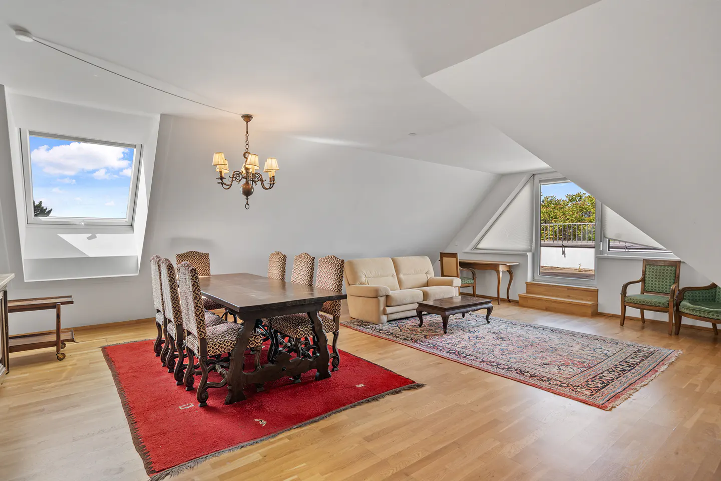 Attic living space with wood floors, white walls, and sloped ceilings. A dining table with chairs sits on a red rug, and a beige sofa is on a patterned rug.
