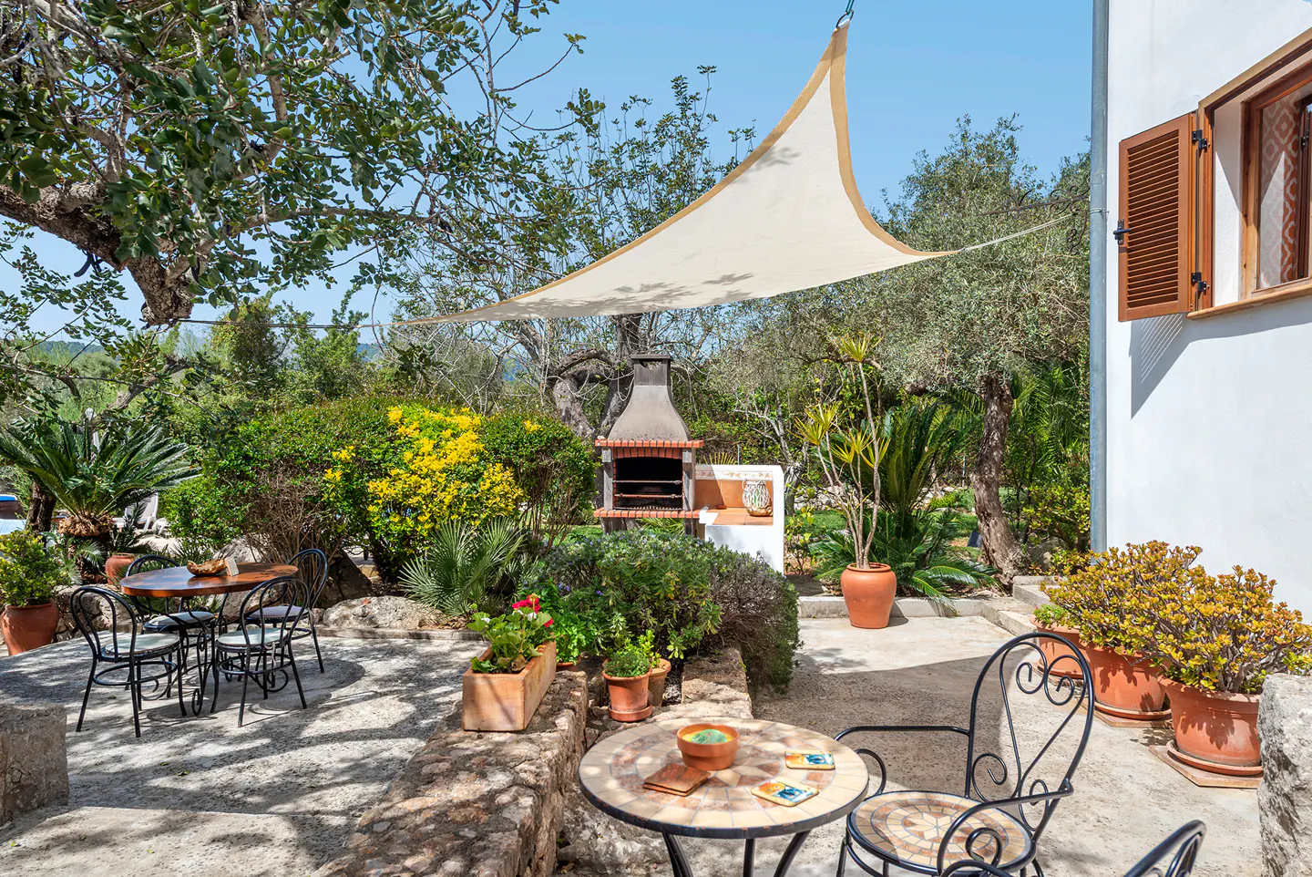 Outdoor patio with stone flooring, tables, chairs, and lush greenery. A beige sunshade is overhead, and a brick barbecue is in the background.