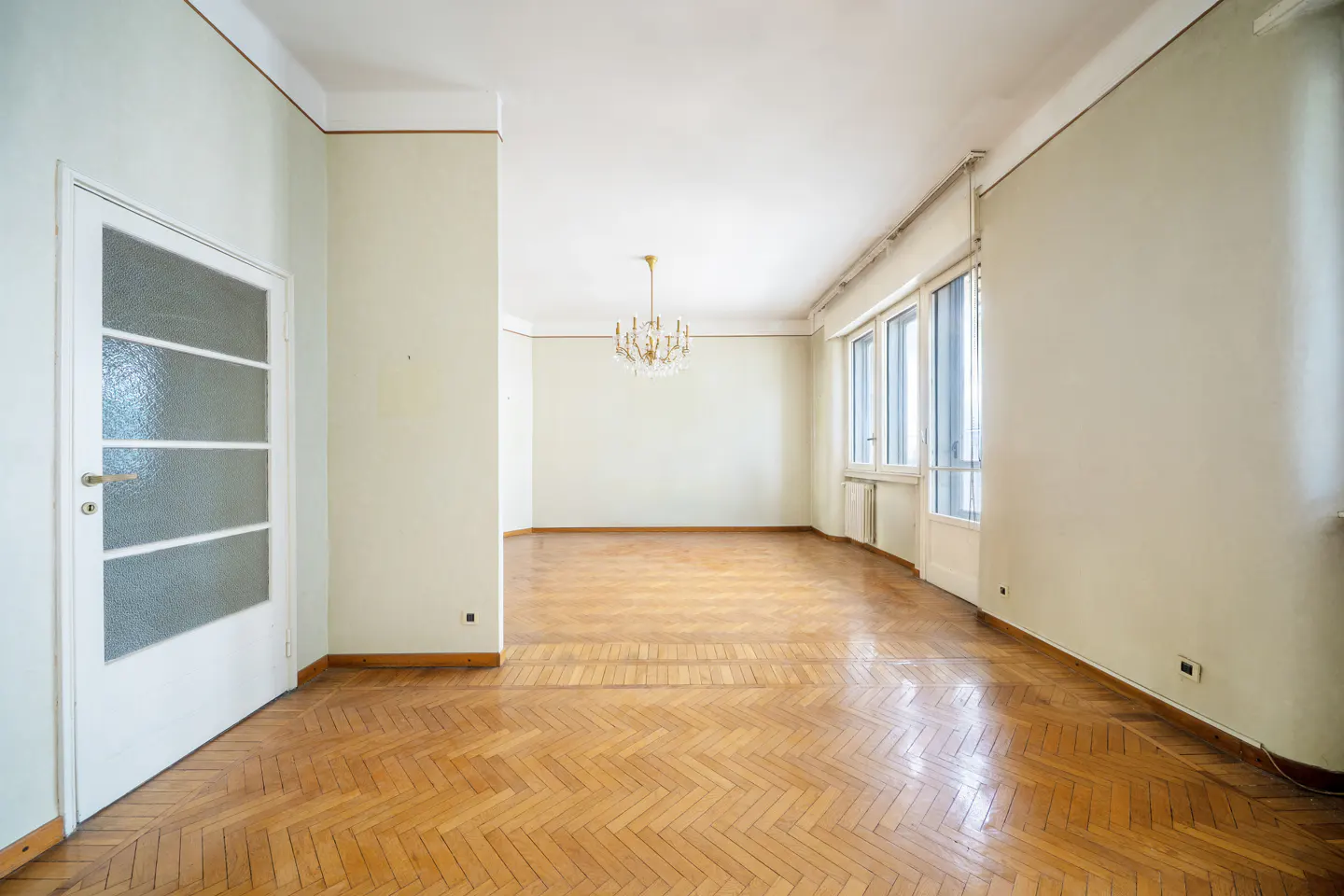 Empty room with herringbone wood floors, white walls, and a crystal chandelier. Windows line one wall. A white door is on the left.