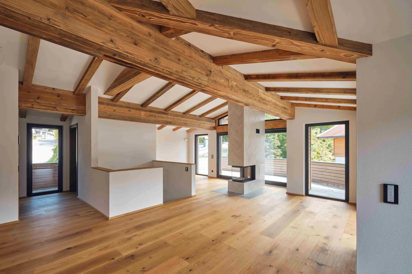 Bright, modern living room with exposed wood beams, light wood floors, and a stone fireplace. Large windows offer outdoor views.