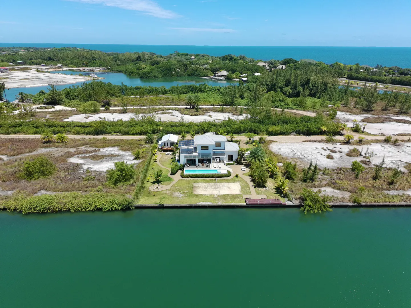 Aerial view of a modern white house with a pool, dock, and volleyball court on a green canal, with ocean and trees in the background.