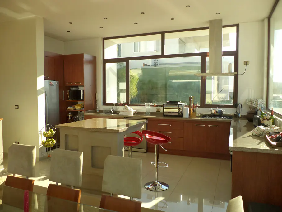 Bright, modern kitchen with brown cabinets, island with red stools, and large windows overlooking greenery.