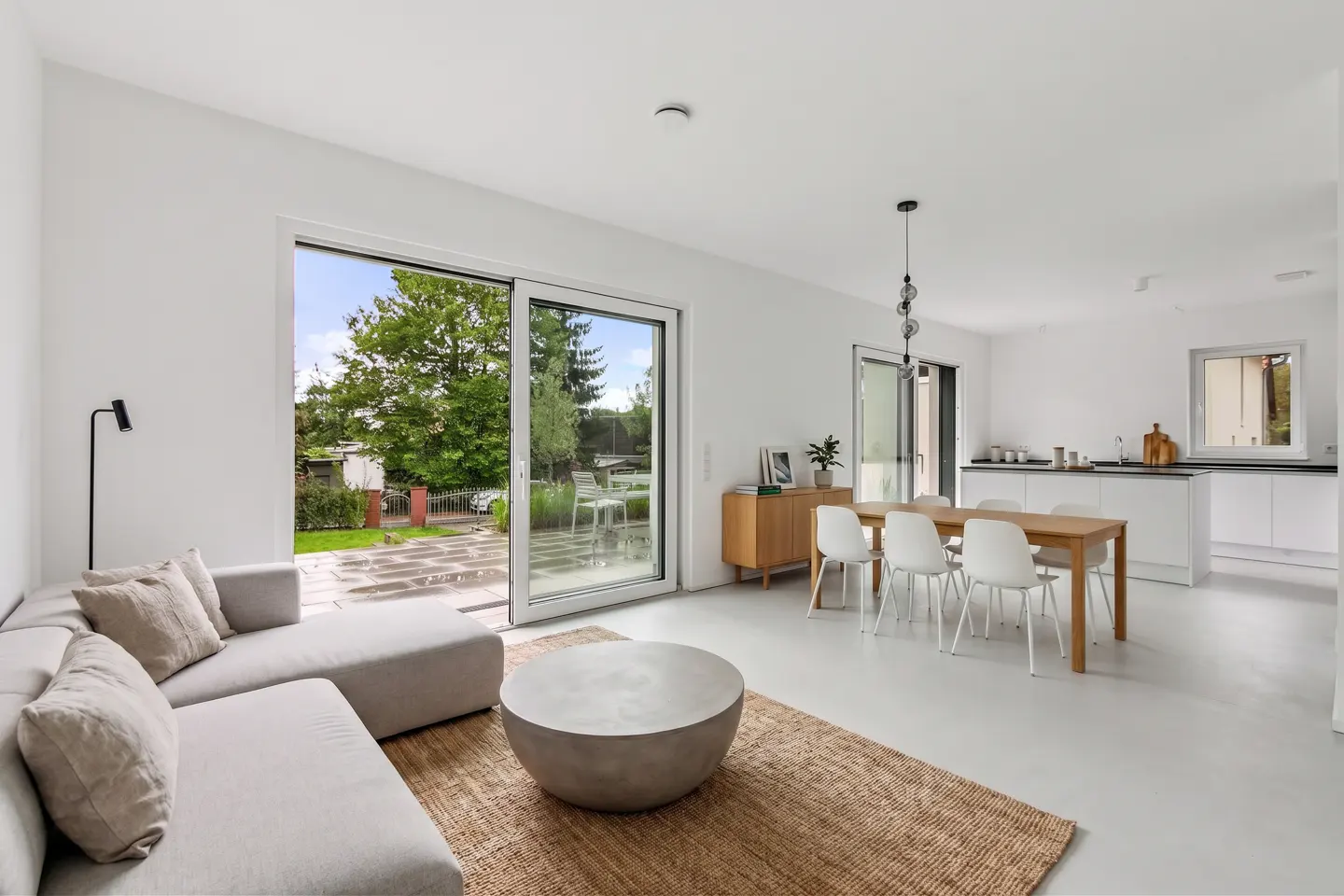 Bright, modern living room with a gray sofa, round coffee table on a jute rug, and a dining table with white chairs. Sliding glass doors open to a green backyard.