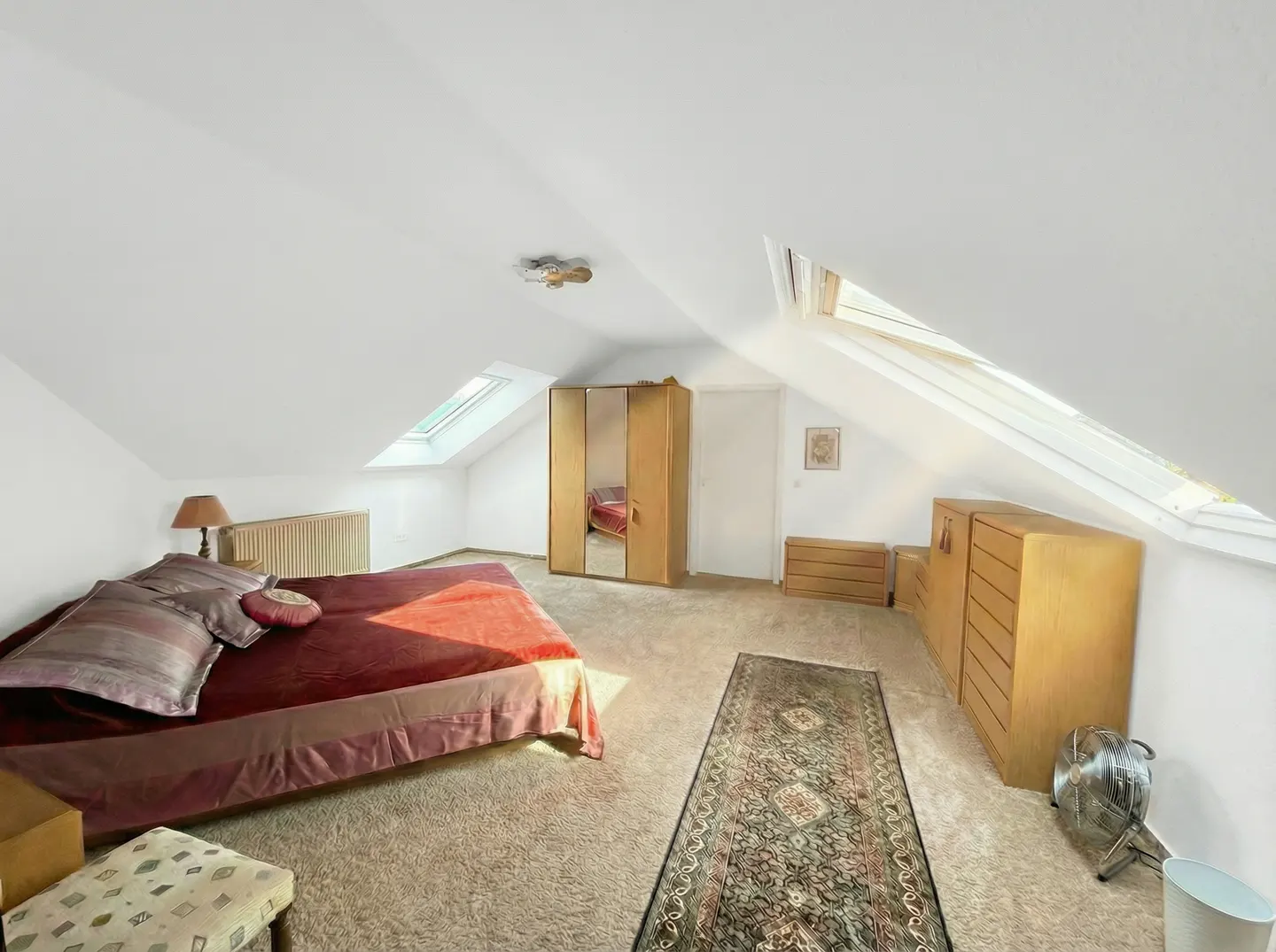 Attic bedroom with white walls, carpet, and skylights. A bed with red bedding, a wardrobe, and wooden dressers furnish the room.