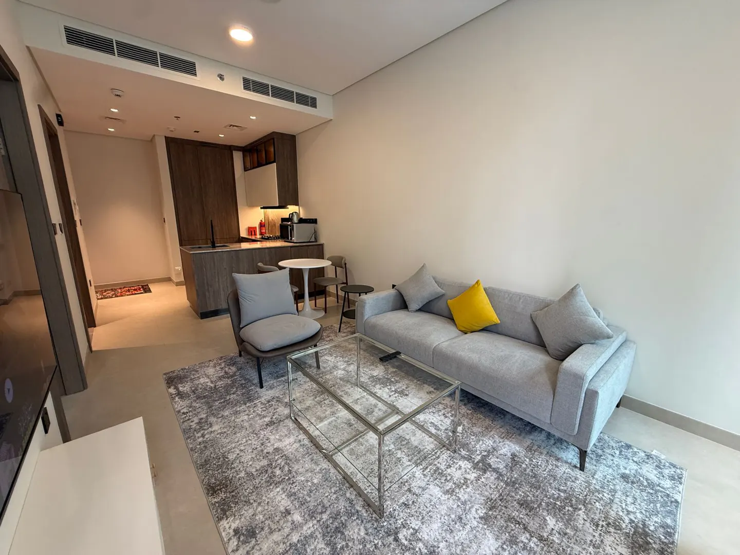 A modern apartment interior with a gray sofa, yellow pillow, glass table, and open kitchen.